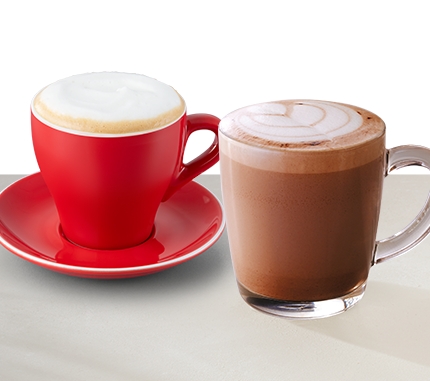 Two hot beverages including a cappuccino in a red cup with saucer and a creamy hot chocolate in a clear glass mug
